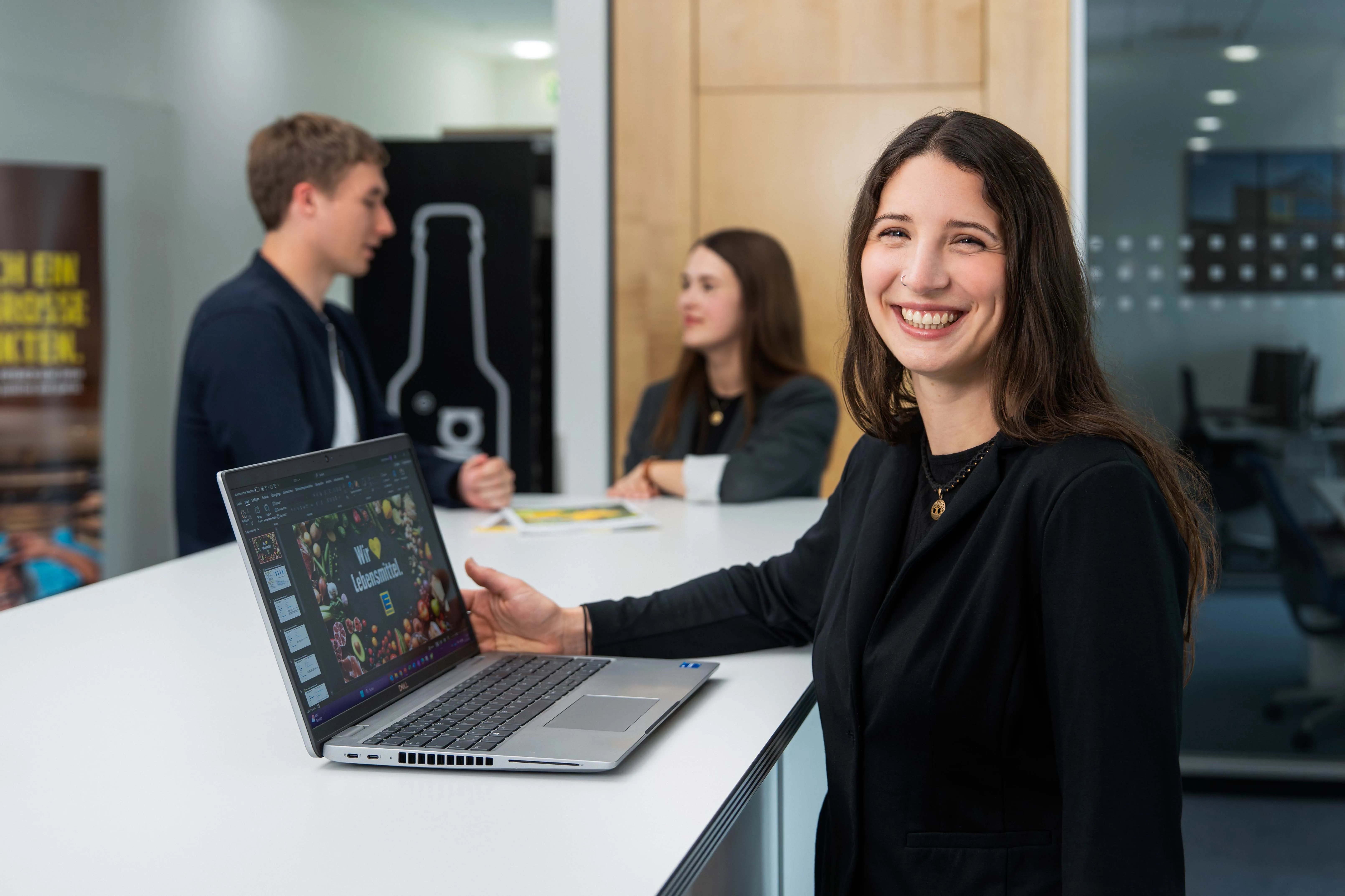 Ein lächelnder Mann schreibt an ein Whiteboard, während Kolleg:innen im Hintergrund an einem Tisch arbeiten.