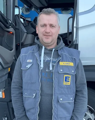 Ein EDEKA-Lkw-Fahrer steht freundlich vor seinem Lkw und lädt Dich ein, Teil des Teams im Einzelhandel zu werden.