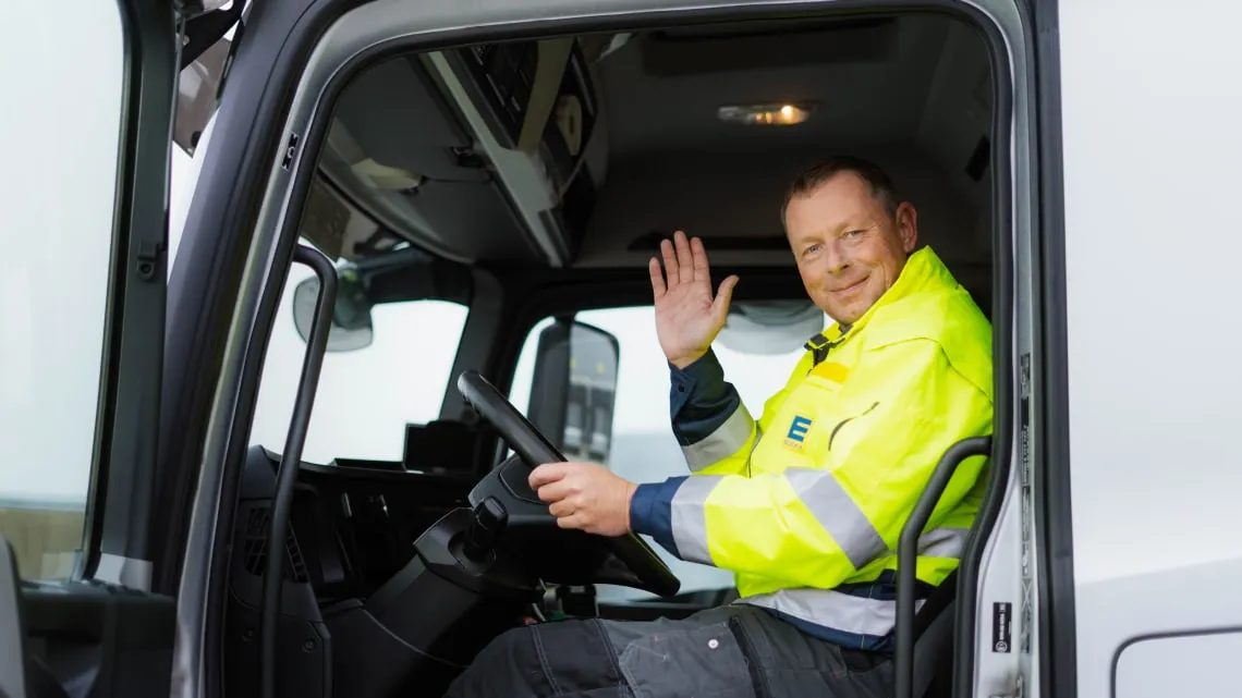 Ein LKW-Fahrer im EDEKA-Outfit sitzt im Fahrersitz und winkt dir freundlich aus dem Führerhaus zu.
