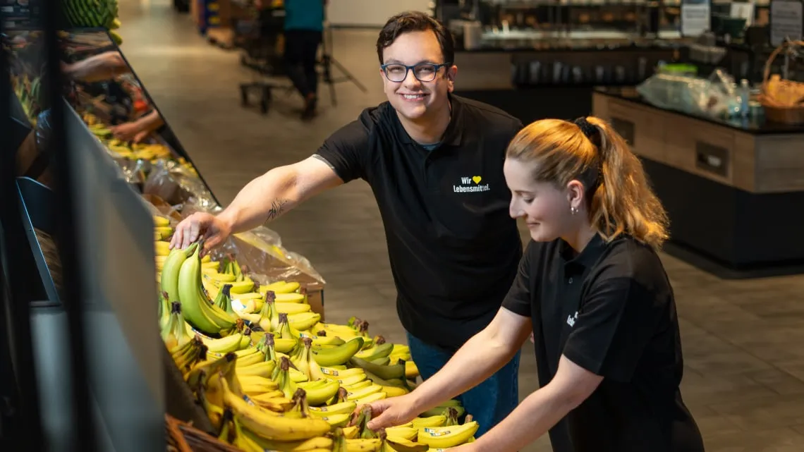 Zwei junge Mitarbeitende sortieren Bananen im EDEKA-Markt und lächeln, während sie im Obstbereich arbeiten.