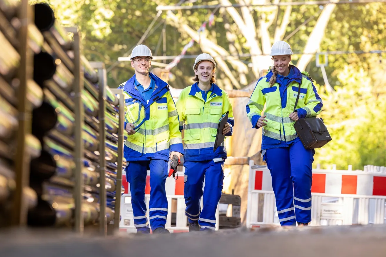 Drei Mitarbeiter von Emscher Lippe Energie in Schutzkleidung und Helmen gehen lachend auf einer Baustelle.