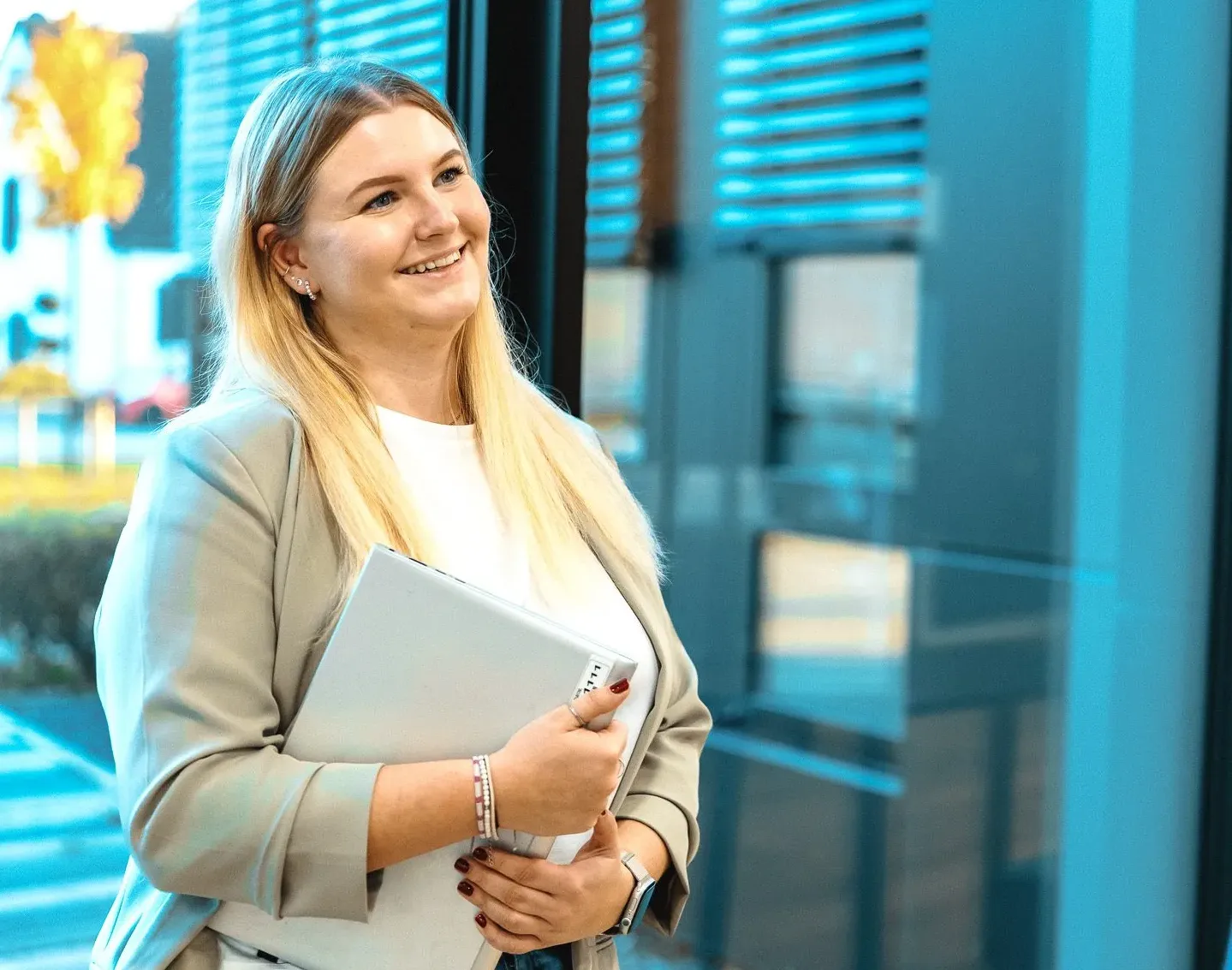 Eine lächelnde Frau in Businesskleidung hält einen Laptop in einem modernen Bürogebäude.