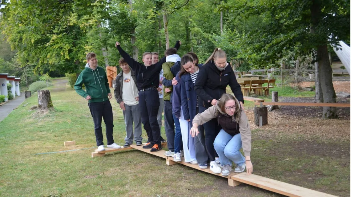 Junge Erwachsene beim Outdoor-Teamtraining balancieren gemeinsam auf Holzbalken, um Teamgeist bei Evonik zu stärken.