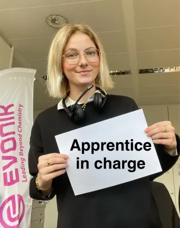 Junge Frau im Büro hält ein Schild mit „Apprentice in charge“ vor einem Evonik-Banner und lächelt in die Kamera.