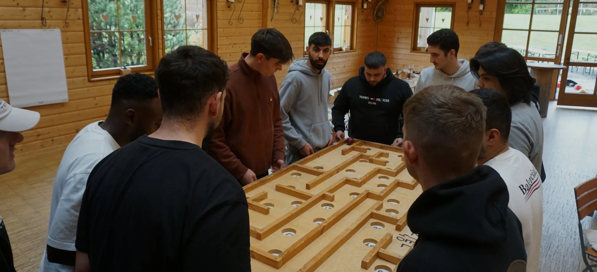 Eine Gruppe von Menschen konzentriert sich auf ein Holzspielbrett in einem Raum mit Holzwänden und Fenstern.