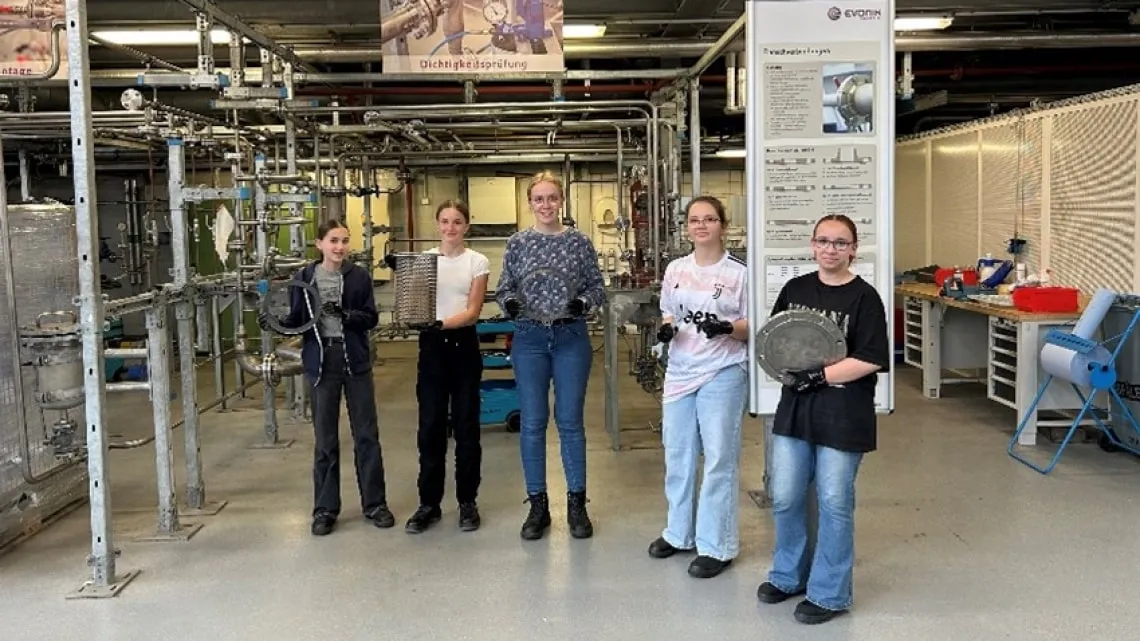Fünf junge Frauen stehen in einer Evonik-Werkshalle und halten technische Bauteile aus dem Energie- und Versorgungsbereich.