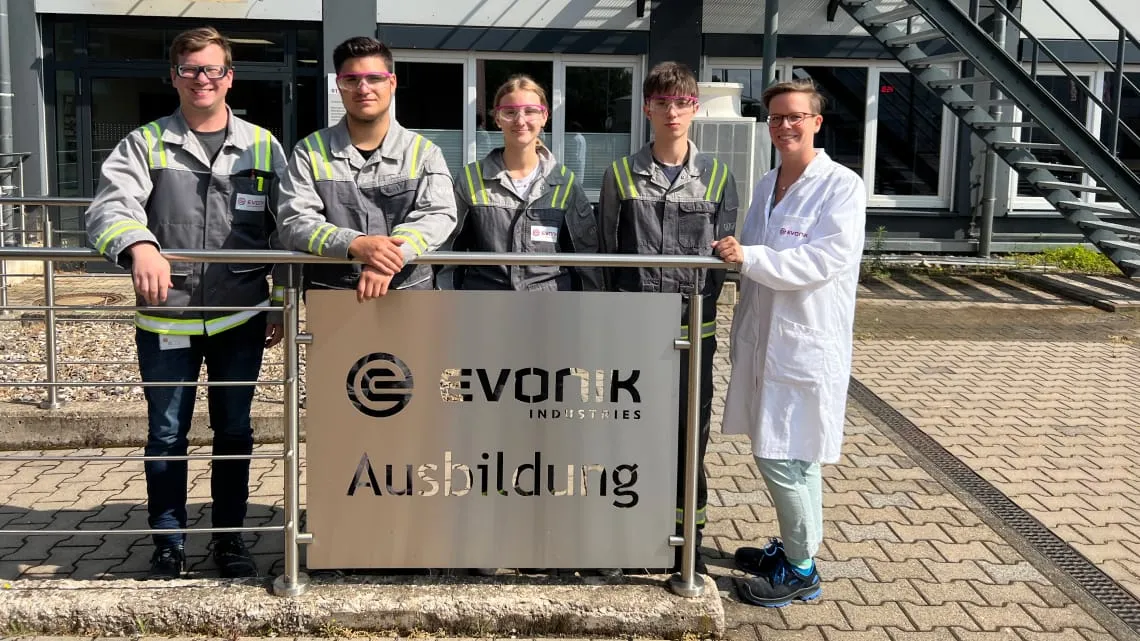 Vier Auszubildende und eine Ausbilderin von Evonik stehen gemeinsam vor einem Schild mit der Aufschrift „Ausbildung“.