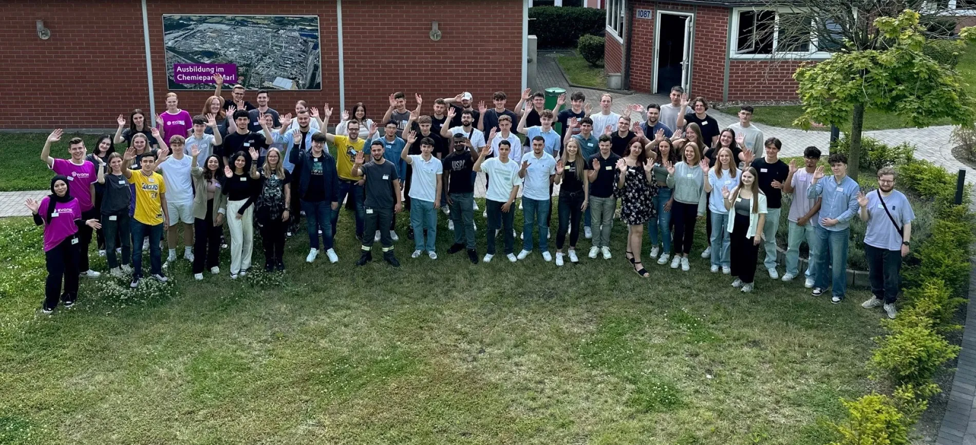 Gruppe junger Menschen vor einem Gebäude im Chemiepark Marl, die gemeinsam in die Kamera winken. Ideal für deine Karriere bei Evonik.