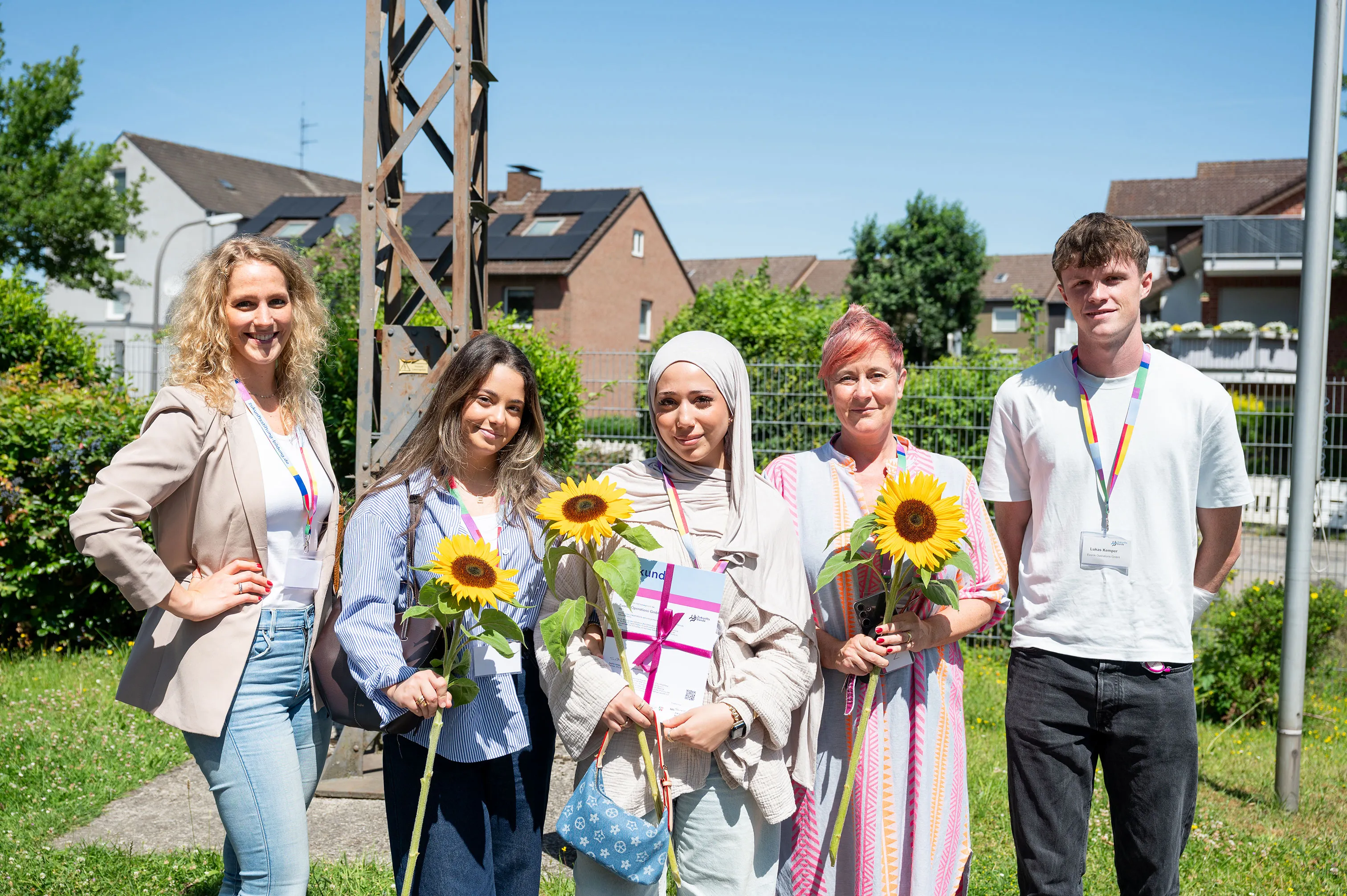 Eine Gruppe von Menschen hält Zertifikate und Sonnenblumen vor einem Gebäude. Sie lächeln in die Kamera.
