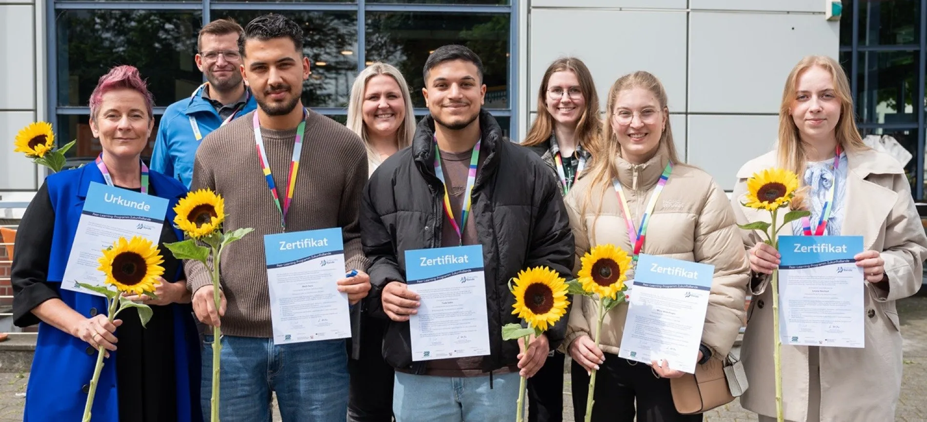 Eine Gruppe von Menschen hält Zertifikate und Sonnenblumen vor einem Gebäude. Sie lächeln in die Kamera.