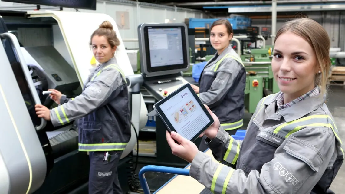 Drei junge Frauen in Arbeitskleidung arbeiten bei Evonik an Maschinen, eine hält ein Tablet und lächelt dich an.
