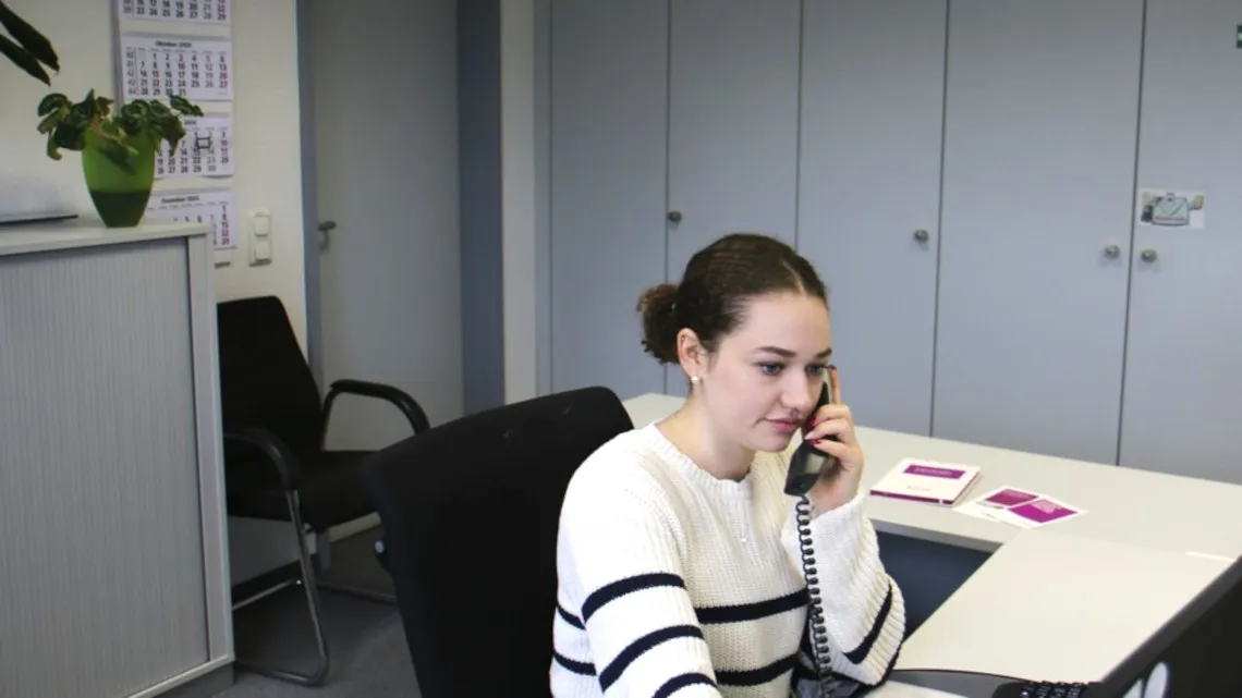 Eine Frau im Büro telefoniert und arbeitet am Computer, umgeben von Ordnern.