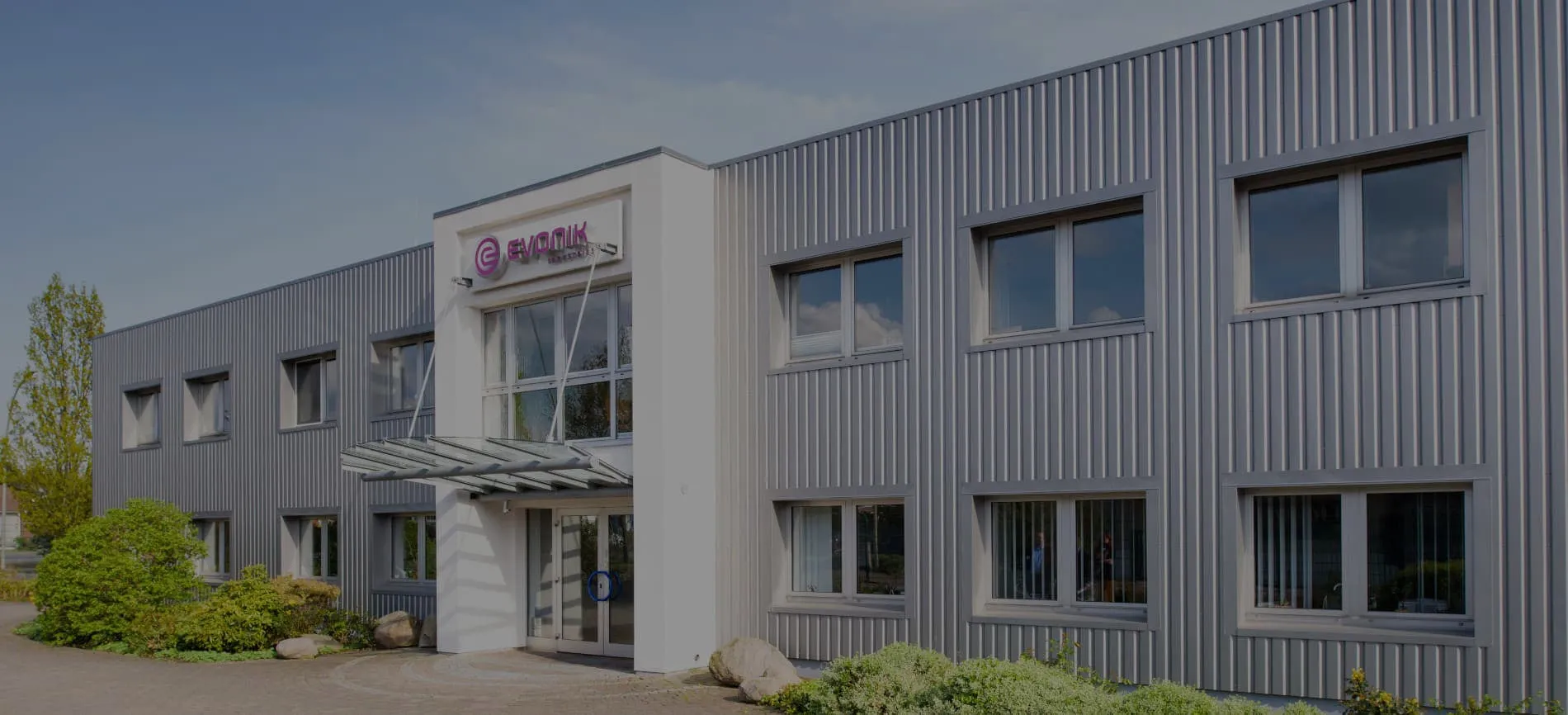 Ein modernes Evonik-Gebäude mit grauer Fassade und Glasdach, umgeben von grüner Bepflanzung, unter blauem Himmel.