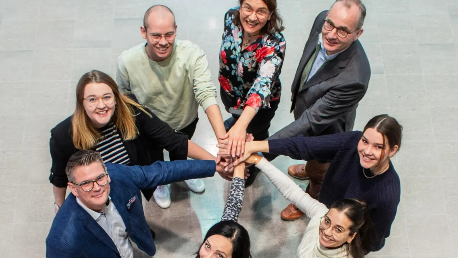 Eine Gruppe von acht FIEGE-Mitarbeitern legt in einem motivierenden Teammeeting die Hände übereinander.