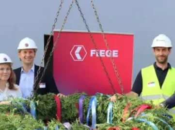 FIEGE-Mitarbeiter beim Richtfest einer Logistikhalle, tragen Helme und stehen vor einem FIEGE-Logo.