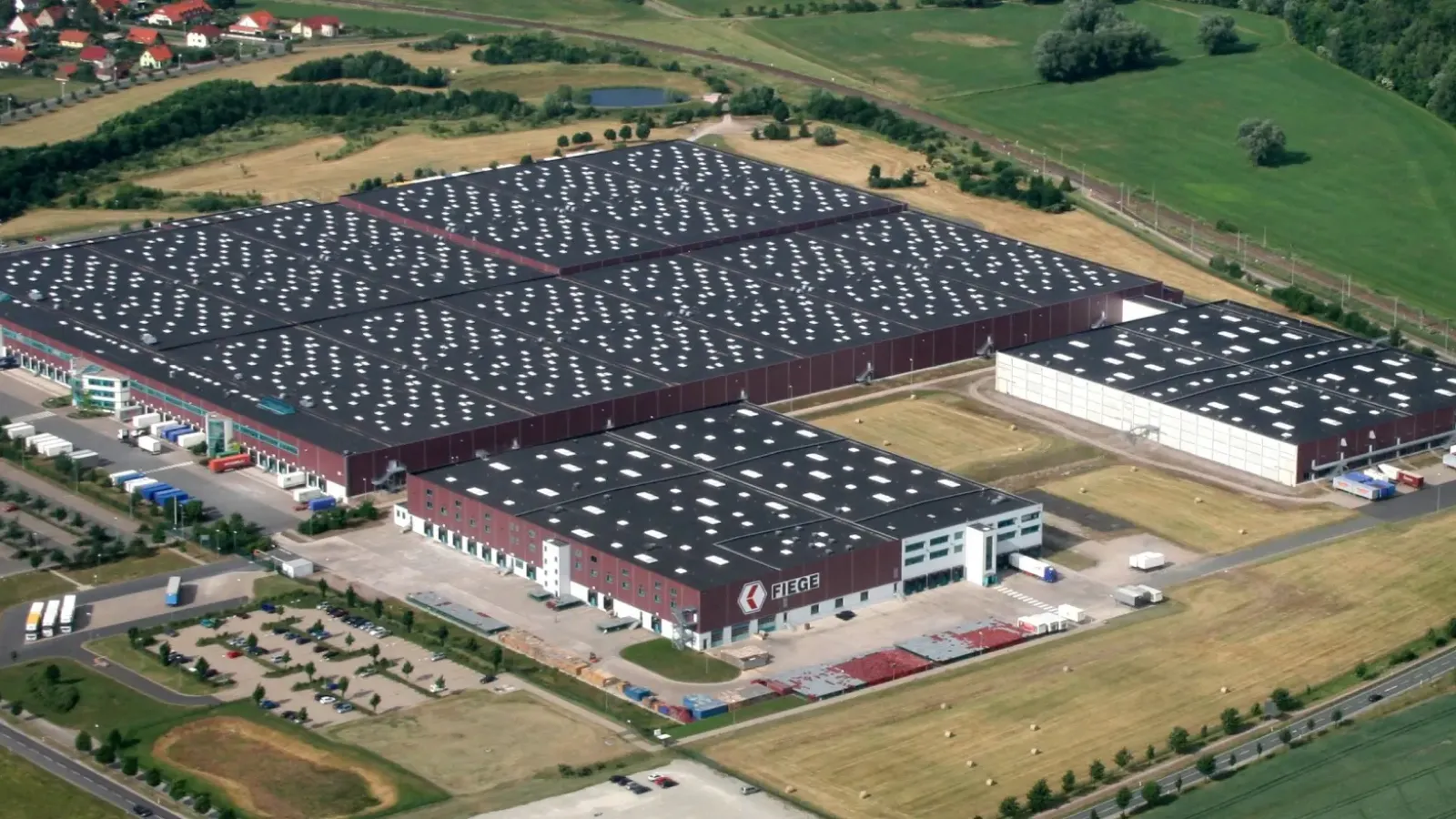 Luftbild eines großen FIEGE-Logistikzentrums mit zahlreichen Lkw, umgeben von Grünflächen und nahe einer Siedlung.