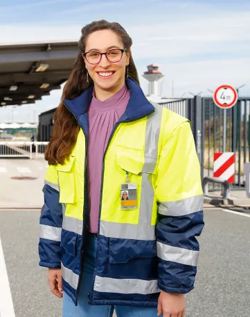 Junge Frau im Sicherheitsbereich des Flughafens mit Warnjacke und Mitarbeiterausweis, bereit für ihren Einsatz bei Fraport.
