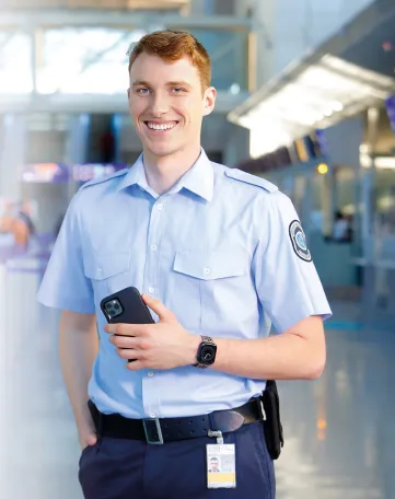 Junger Sicherheitsmitarbeiter am Flughafen lächelt freundlich und hält ein Smartphone, bereit für Deinen Einstieg bei Fraport.