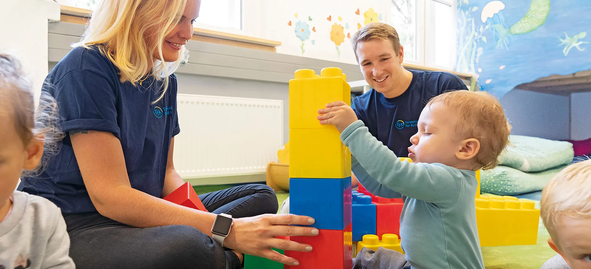 Zwei Mitarbeitende spielen in der betrieblichen Kita von Fraport AG mit kleinen Kindern und bunten Bauklötzen.
