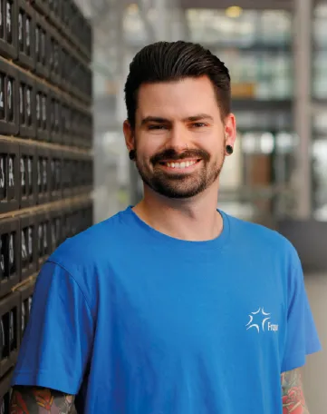 Junger Mitarbeiter im blauen Fraport-T-Shirt lächelt freundlich in moderner Arbeitsumgebung.