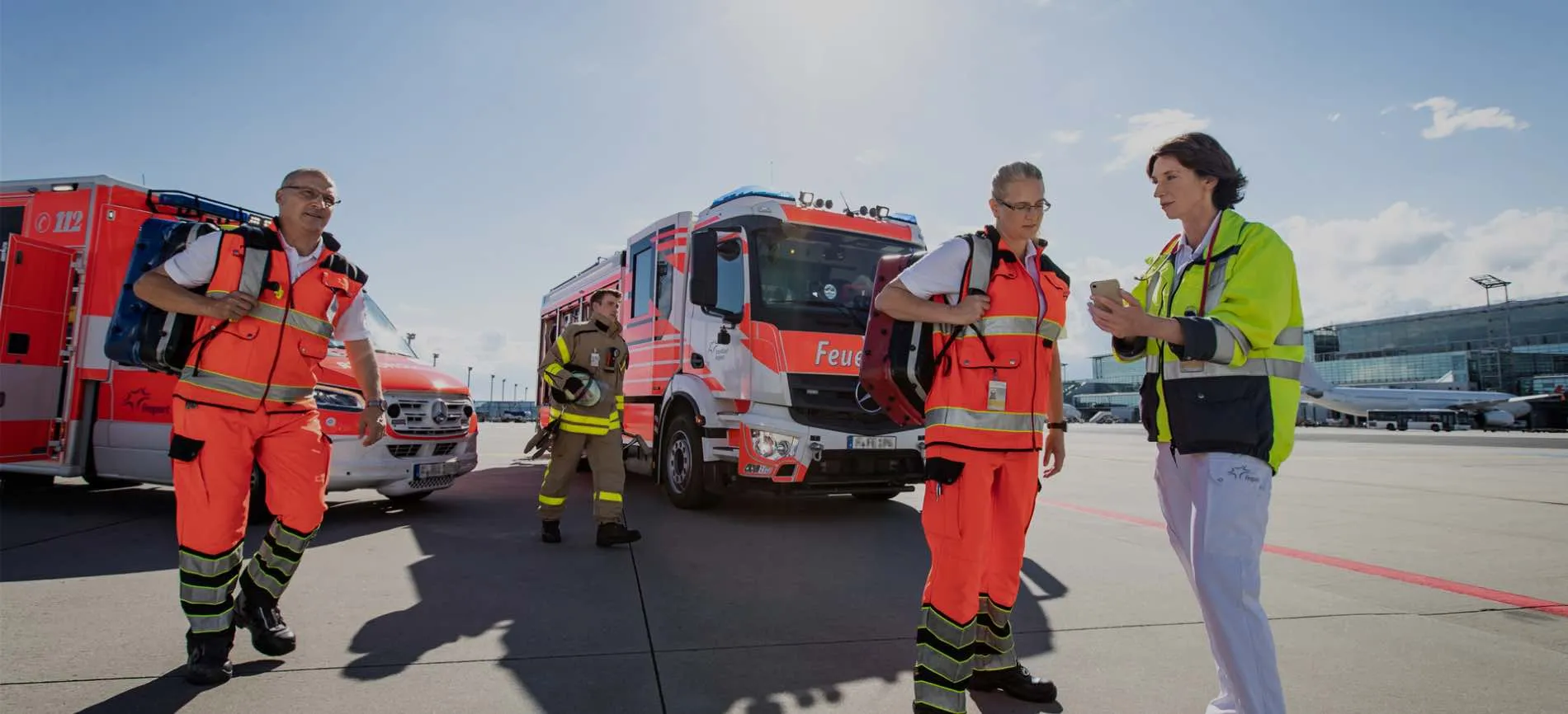 Rettungskräfte und Feuerwehr von Fraport besprechen sich am Flughafen vor Einsatzfahrzeugen im Sonnenschein.