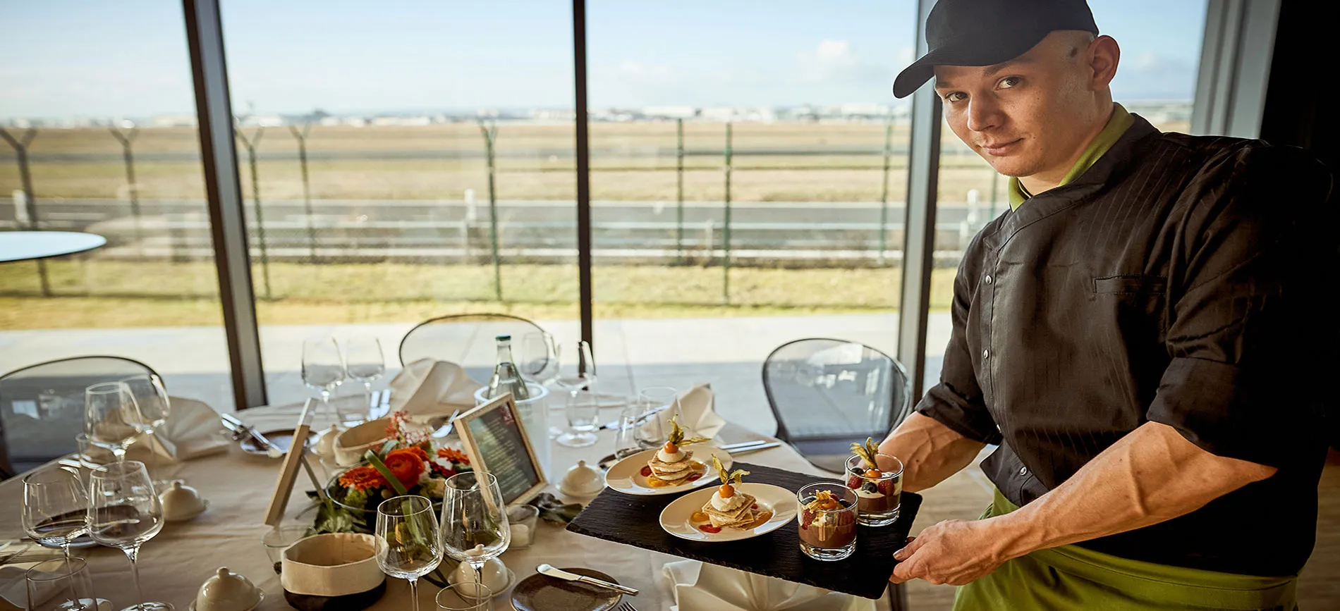 Ein Koch serviert Desserts in einem modern gedeckten Restaurant mit Blick auf das Flughafengelände.