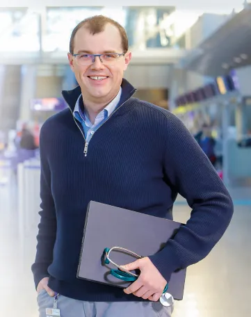 Ein Mitarbeiter von Fraport AG steht mit Laptop und Headset in der Hand freundlich im Flughafenterminal.