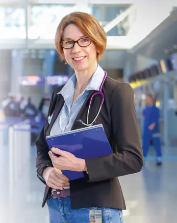 Eine freundliche Mitarbeiterin mit Stethoskop und Aktenmappe steht im Terminal und lädt Dich ein, Teil des Fraport-Teams zu werden.