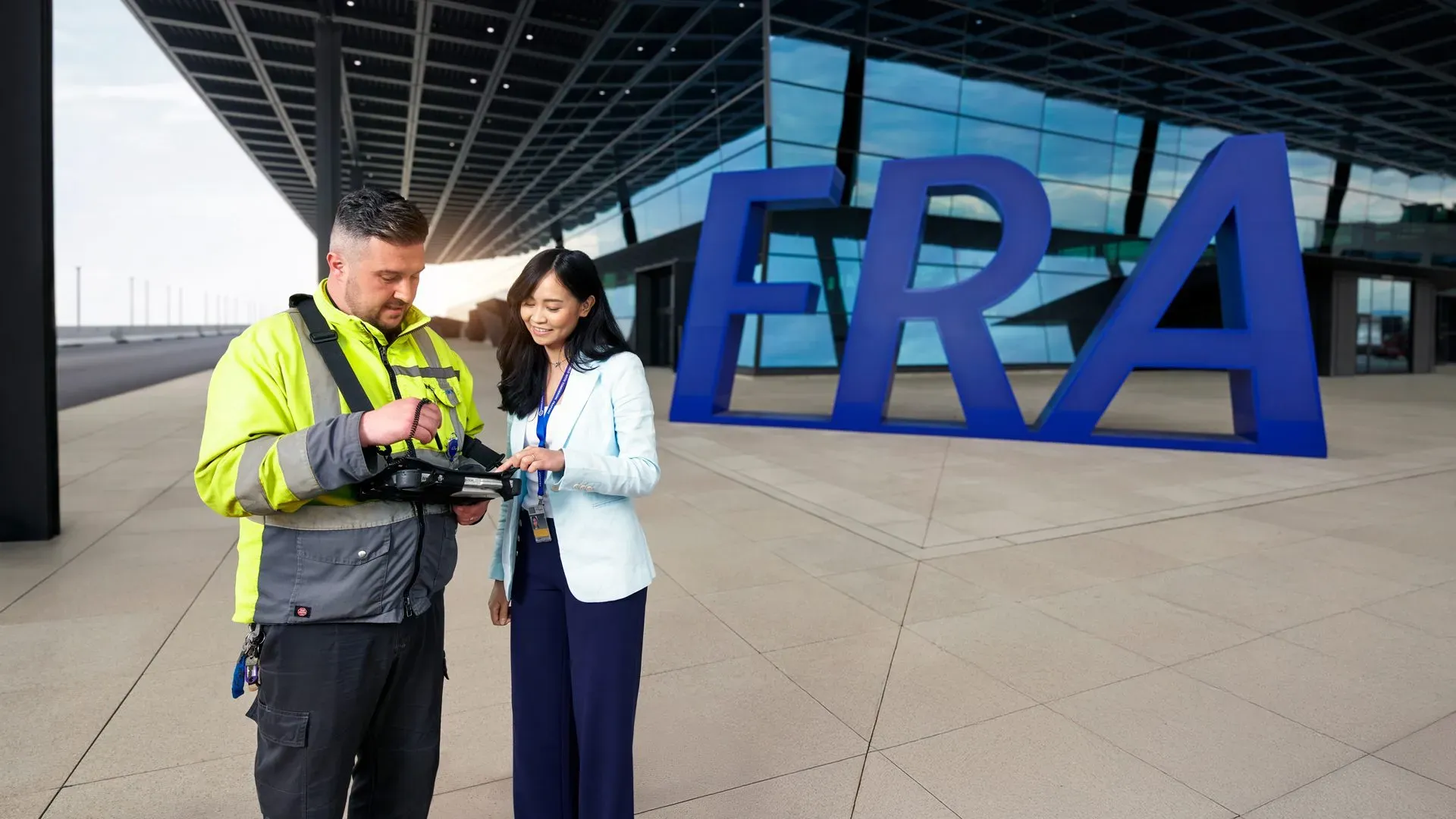 Du siehst eine Fraport-Mitarbeiterin mit Warnweste und Headset lächelnd auf dem Flughafenvorfeld vor einem Flugzeug.