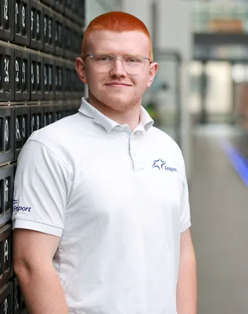 Ein junger Mann im Fraport-Polohemd steht lächelnd im modernen Logistikbereich und lädt Dich zum Mitmachen ein.