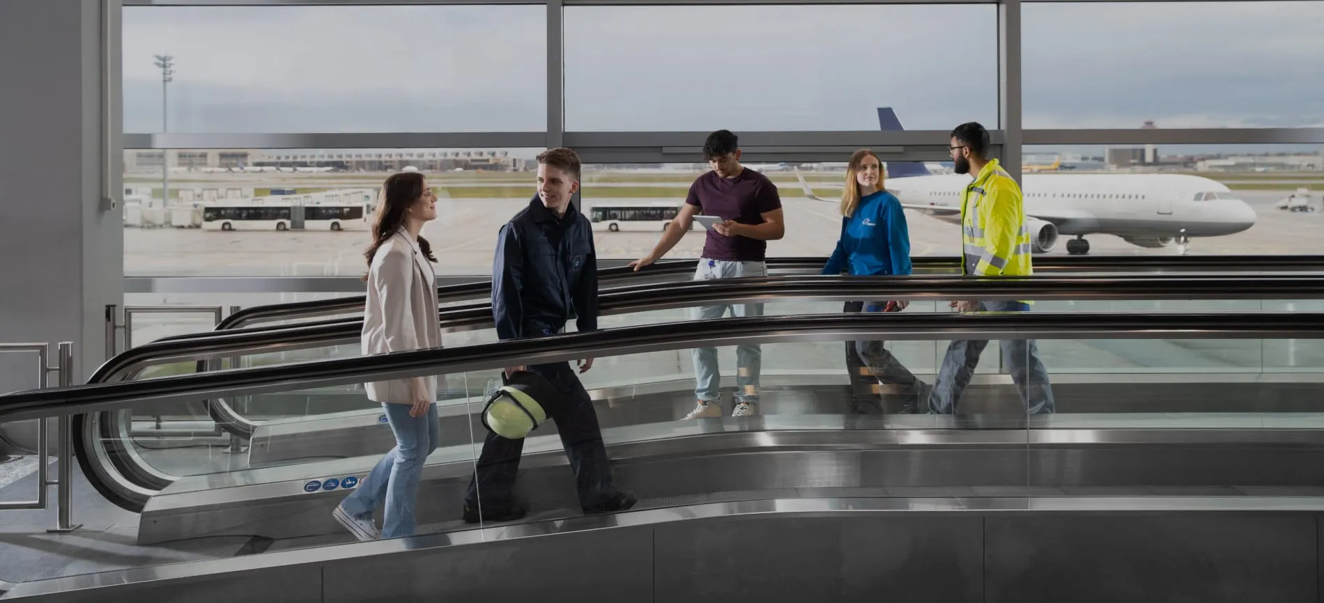 Mehrere junge Menschen in Arbeitskleidung und Freizeitkleidung gehen im Flughafenterminal aneinander vorbei, im Hintergrund ein Flugzeug.