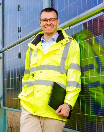 Junger Mitarbeiter mit Fraport-Jacke und Tablet lächelt vor Solarpaneelen – deine Karriere im Transport und Logistik.