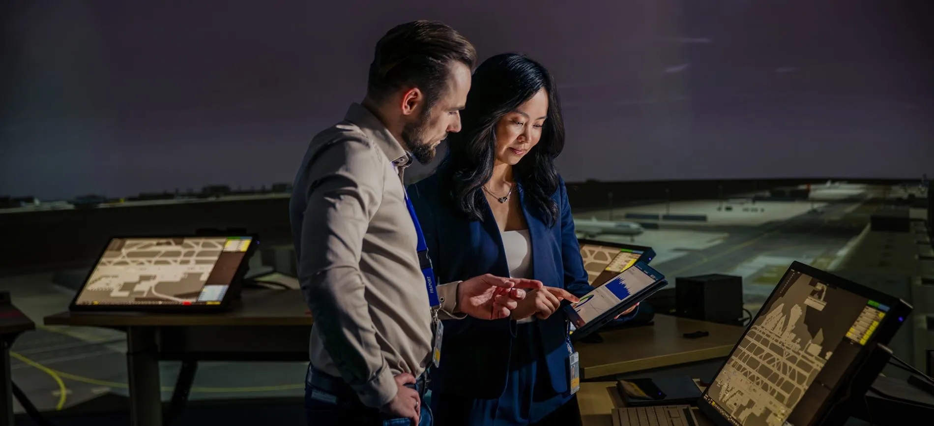 Zwei Mitarbeitende am Flughafen-Arbeitsplatz besprechen Daten auf einem Tablet vor Monitoren mit Flughafenplänen.