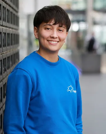 Junge Person im blauen Fraport-Pullover lächelt freundlich in modernem Bürogebäude.