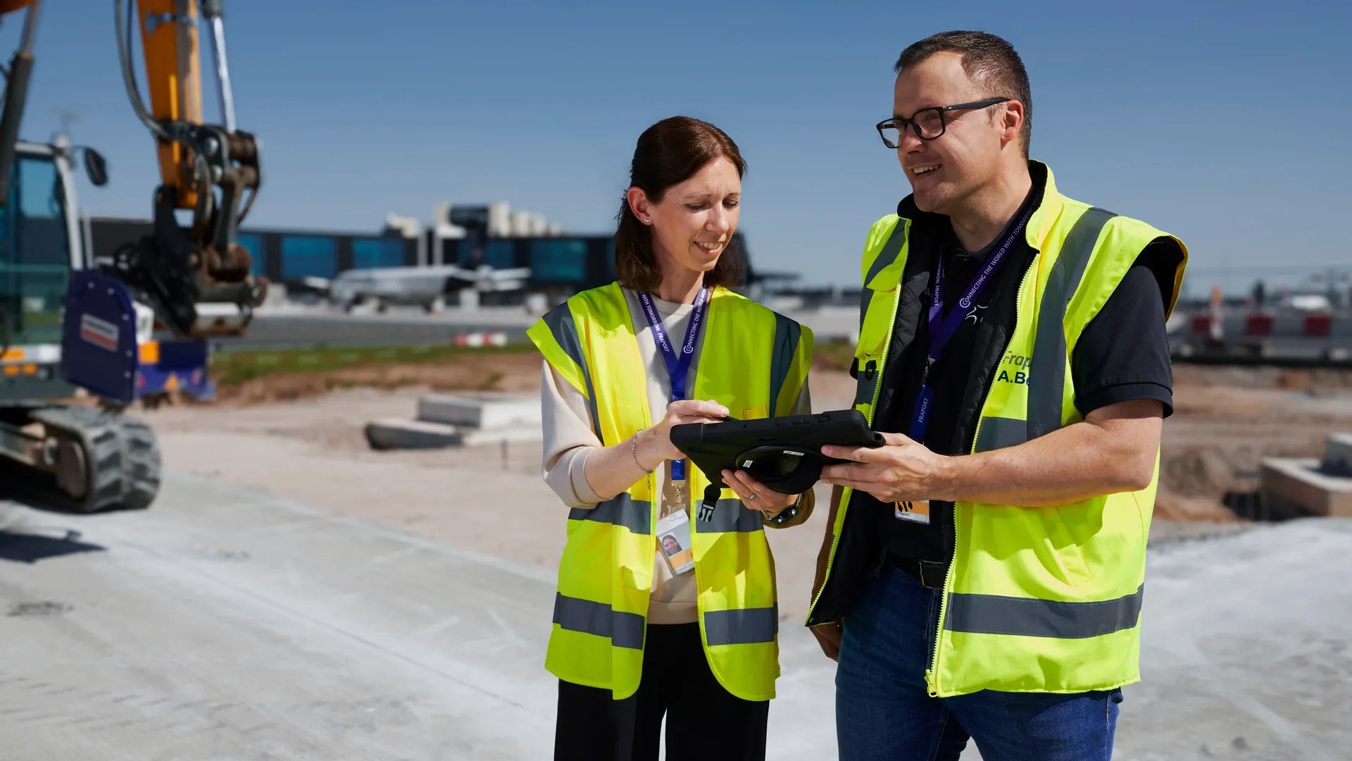 Du siehst ein Kamerateam, das zwei Fraport-Mitarbeitende mit Warnwesten und Helmen am Flughafen interviewt.