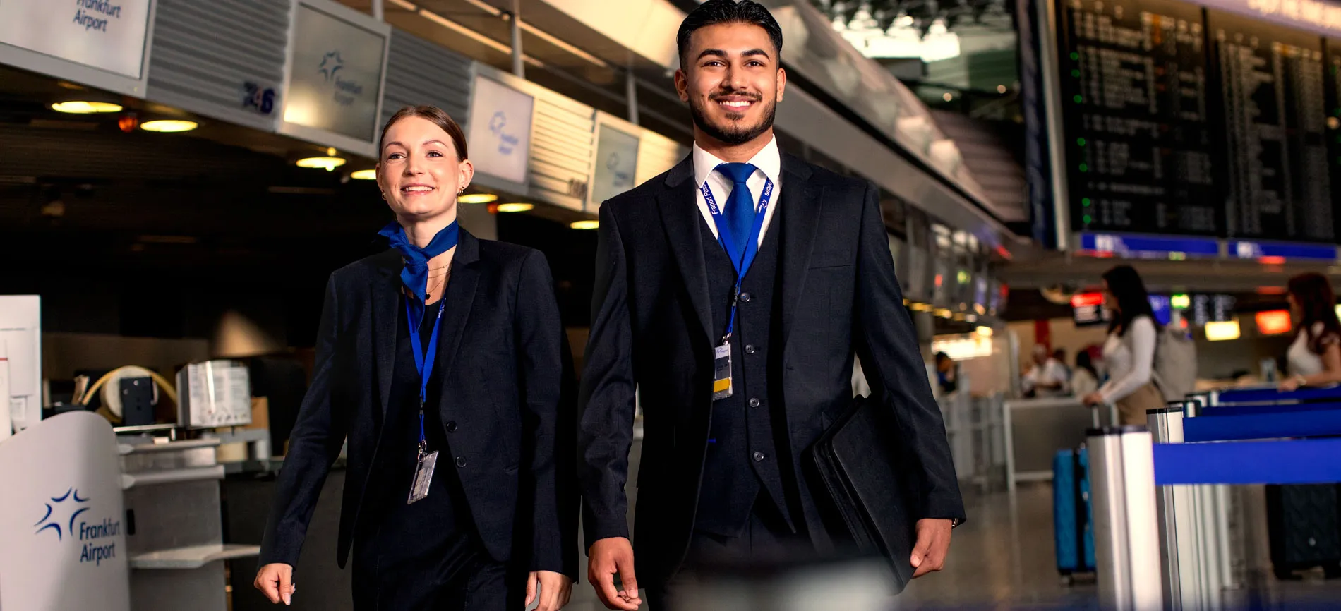 Zwei Fraport-Mitarbeitende in Uniform lächeln und gehen gemeinsam durch den Flughafen Frankfurt.