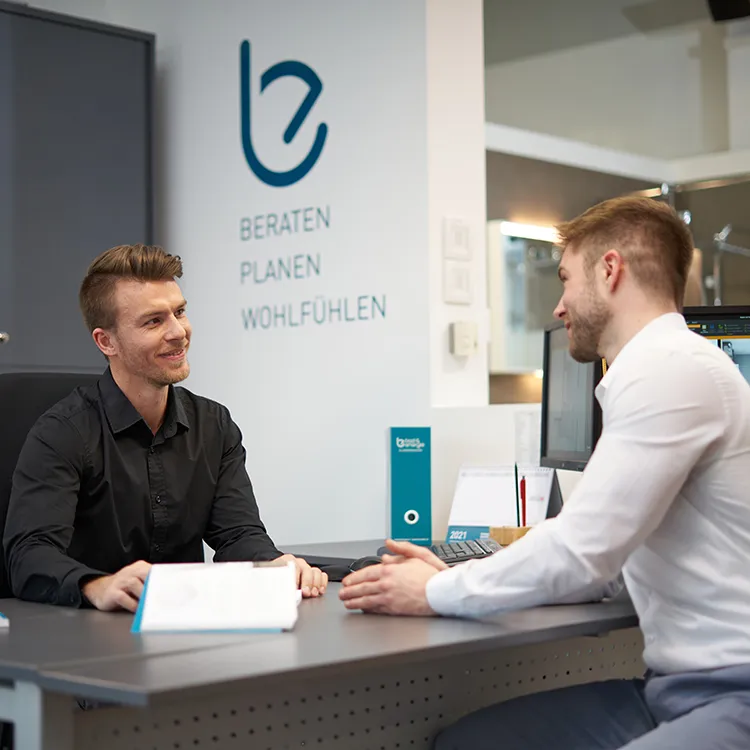 Zwei Männer im Gespräch an einem Schreibtisch in einem modernen Büro der Frauenthal Handel Gruppe.