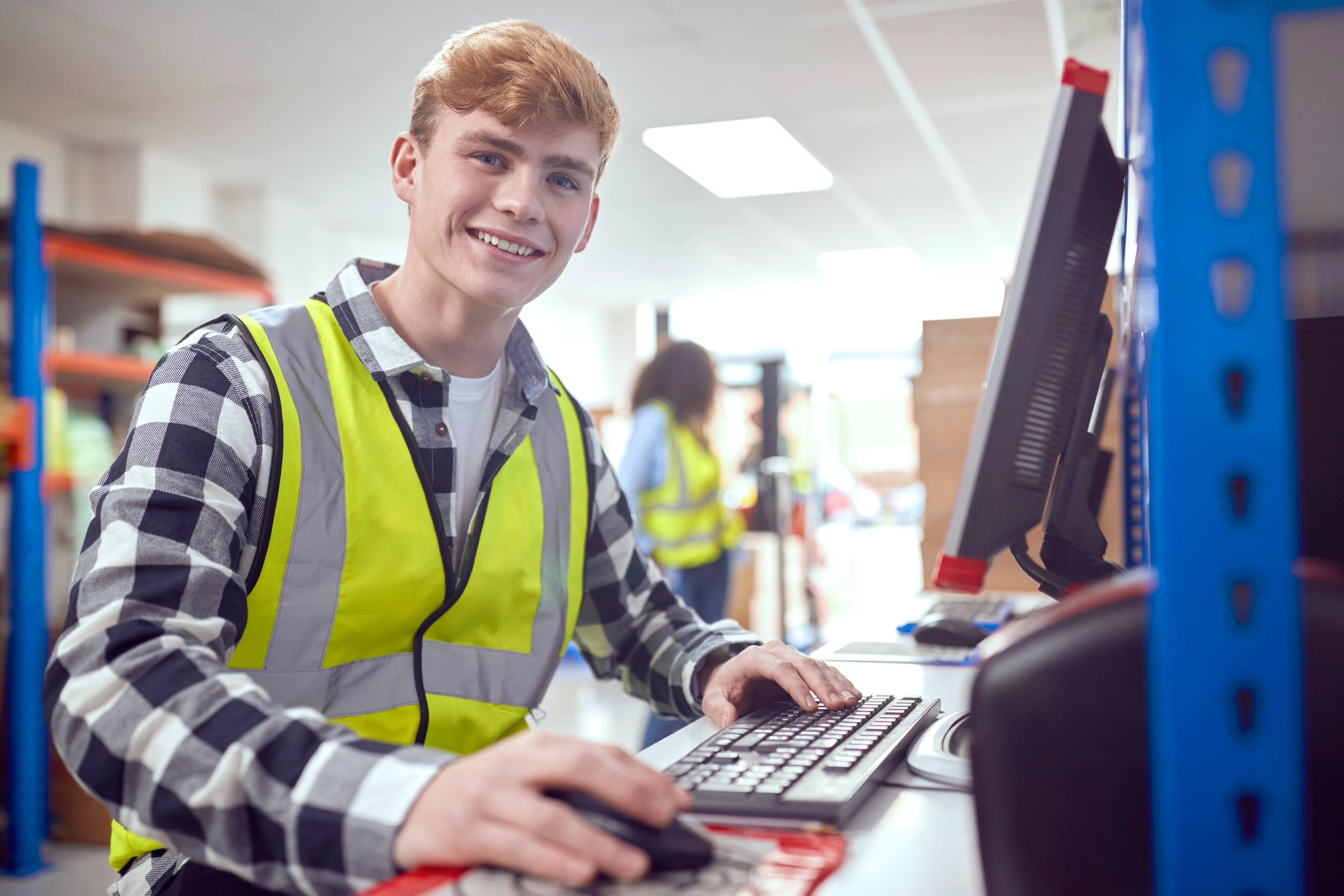 Junger Mitarbeiter mit Warnweste arbeitet am Computer im Lager bei Frauenthal Handel Gruppe und lächelt dich an.