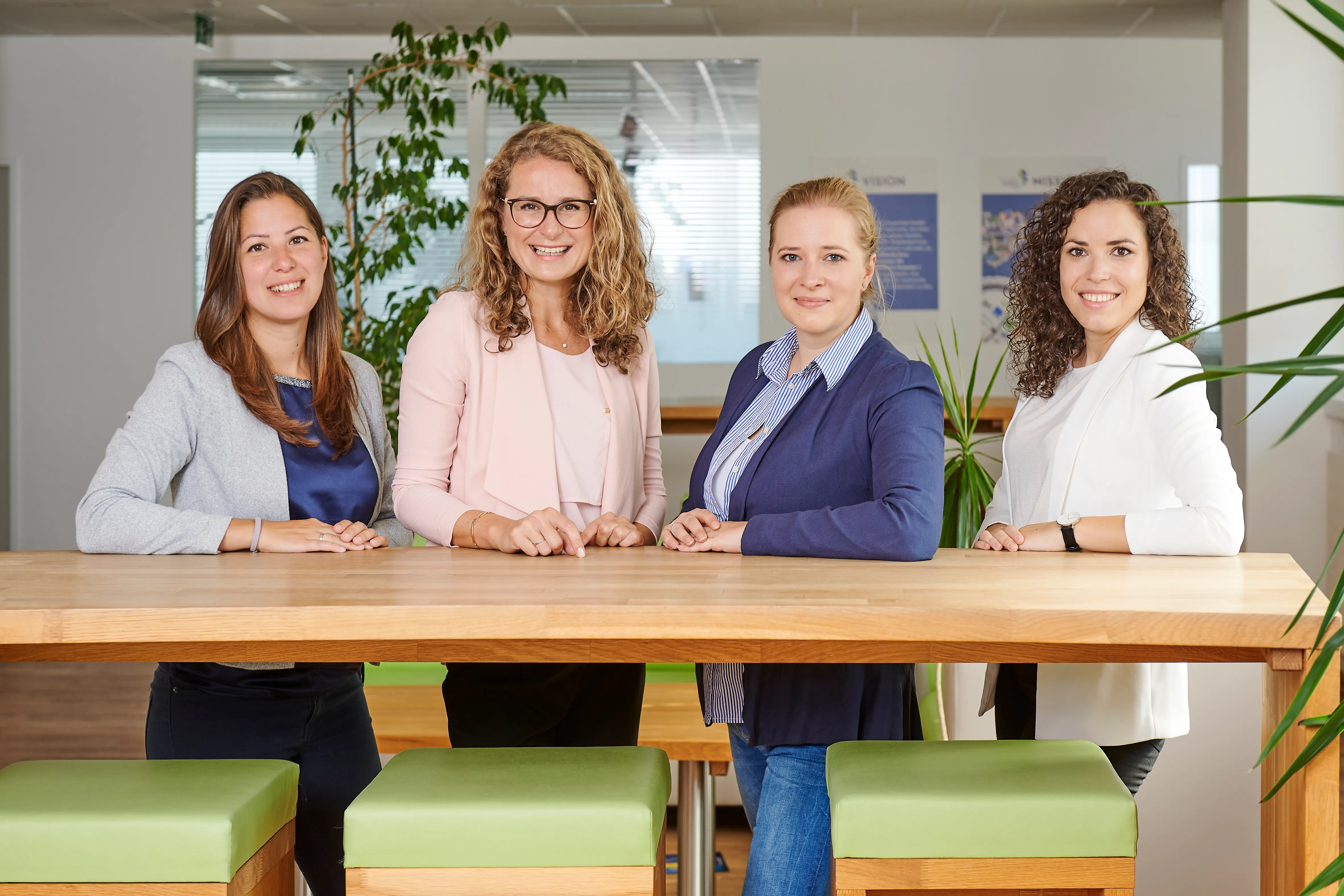 Drei lächelnde Frauen stehen an einem Holztisch in einem modernen Büro bei Frauenthal Handel Gruppe.