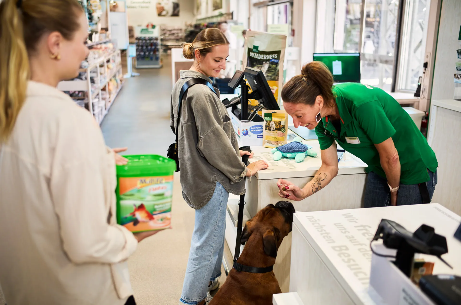 Mitarbeiterin bei Fressnapf berät lächelnd eine Kundin, während sie einem Hund im Geschäft Aufmerksamkeit schenkt.