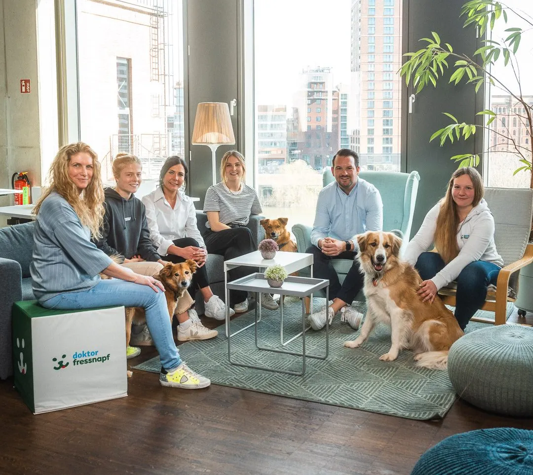 Mitarbeiter von Fressnapf Holding SE sitzen in einem modernen Büro und lächeln, umgeben von Hunden.