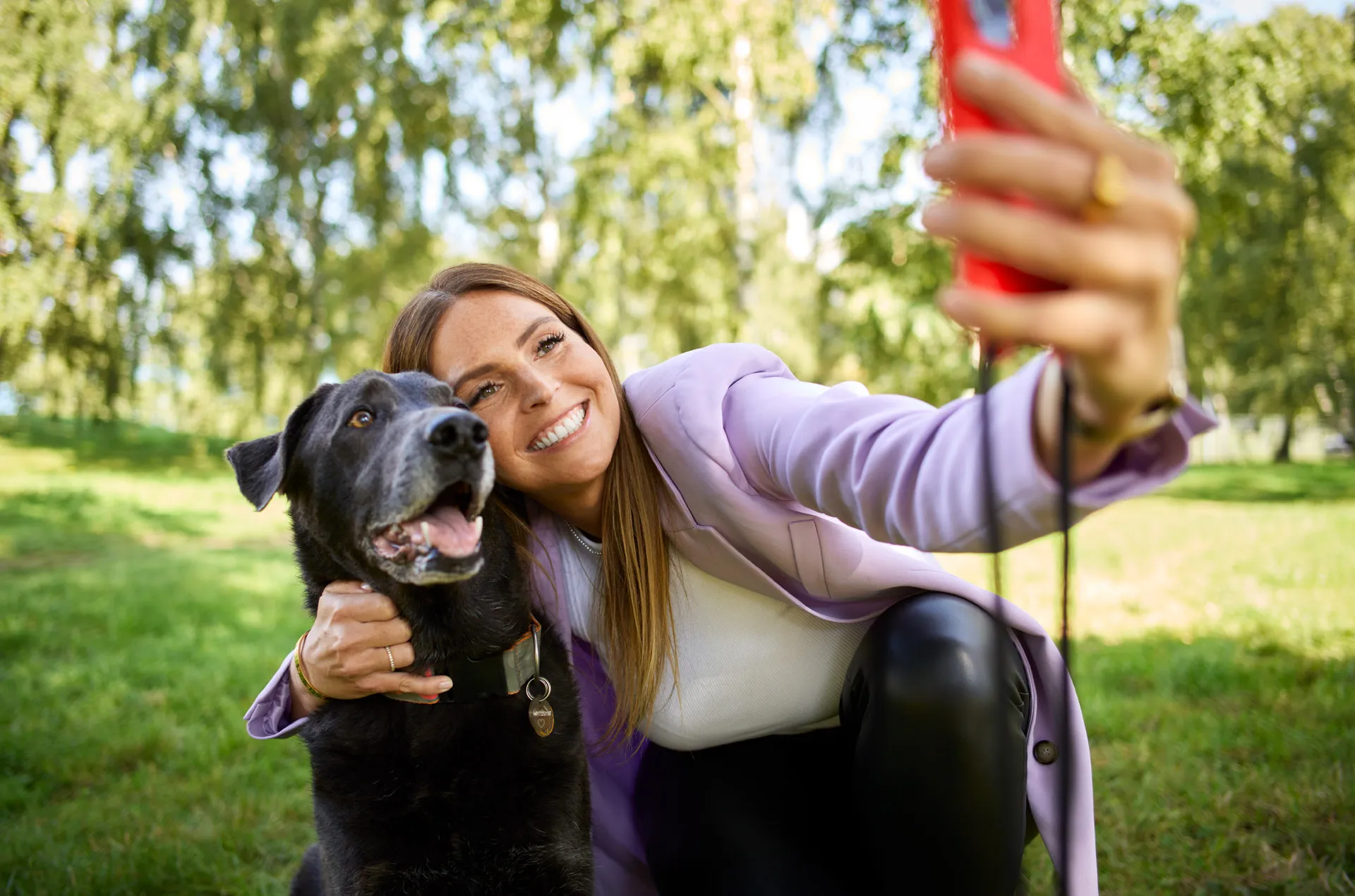 Eine Frau lächelt, während sie im Park ein Selfie mit einem schwarzen Hund macht.