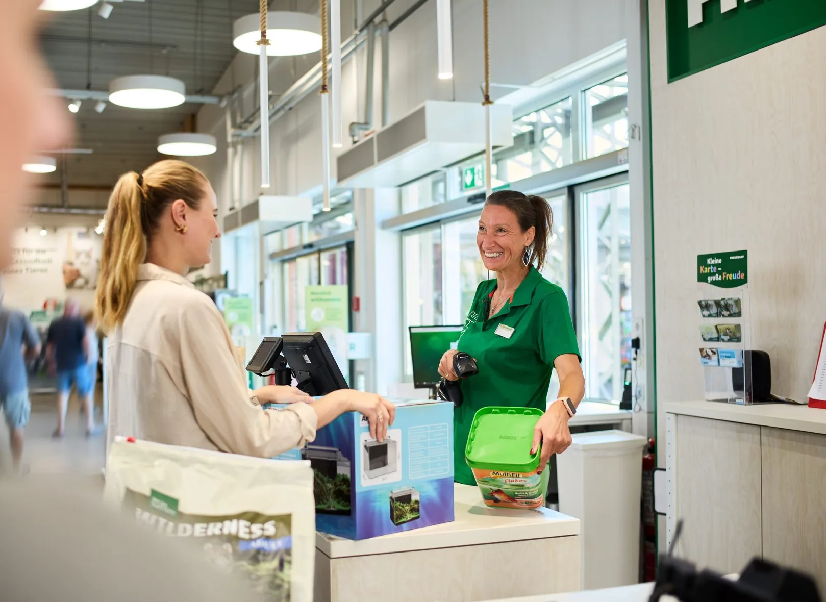 Eine lächelnde Kassiererin im Fressnapf-Geschäft bedient eine Kundin an der Kasse mit einem freundlichen Gespräch.