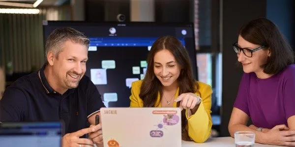 Drei Kollegen bei Fressnapf Holding SE lächeln und arbeiten gemeinsam an einem Laptop in einem modernen Büro.