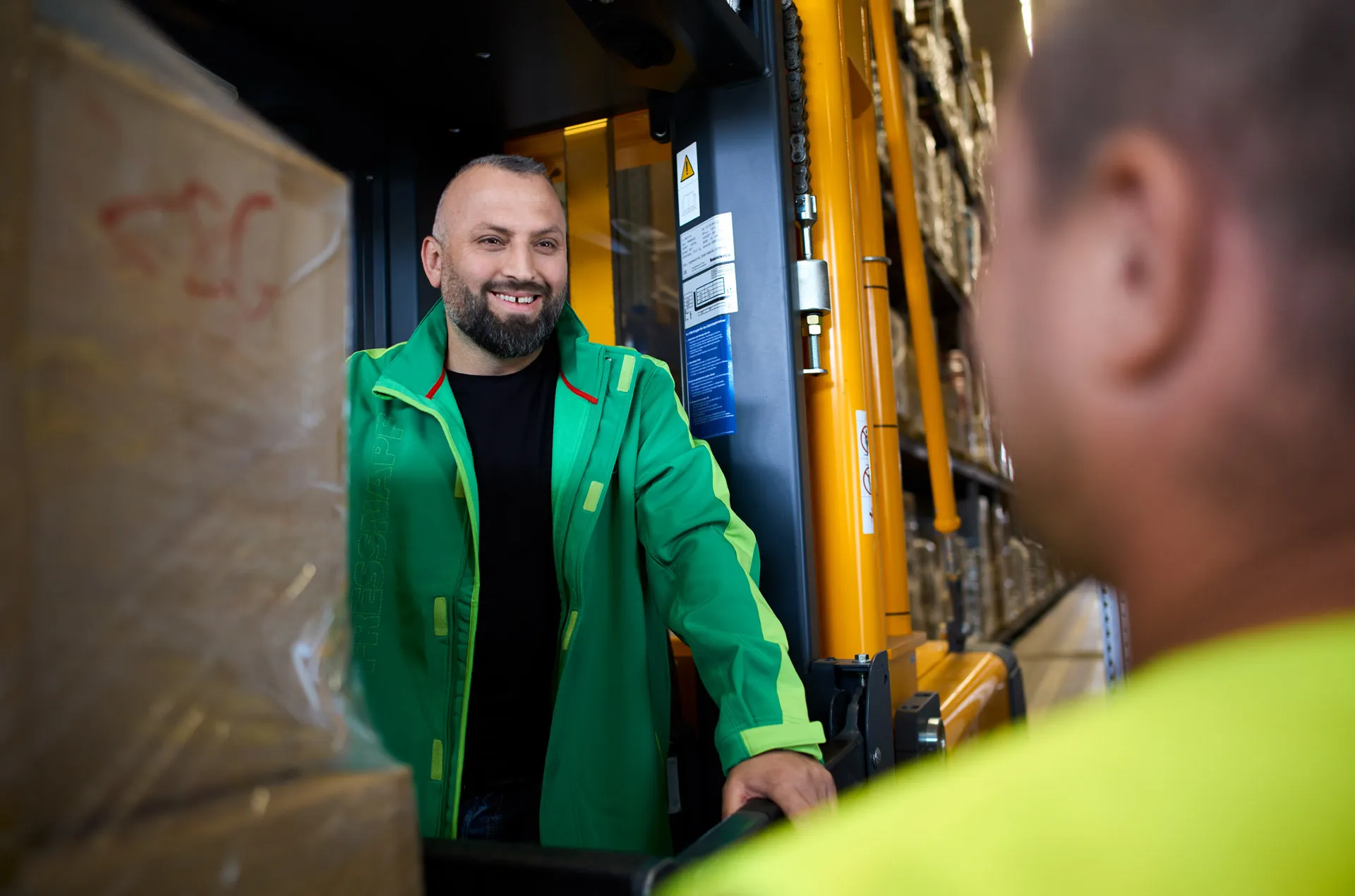 Ein Lagerarbeiter in grüner Arbeitskleidung spricht lächelnd mit einem Kollegen in einem Lager von Fressnapf Holding SE.