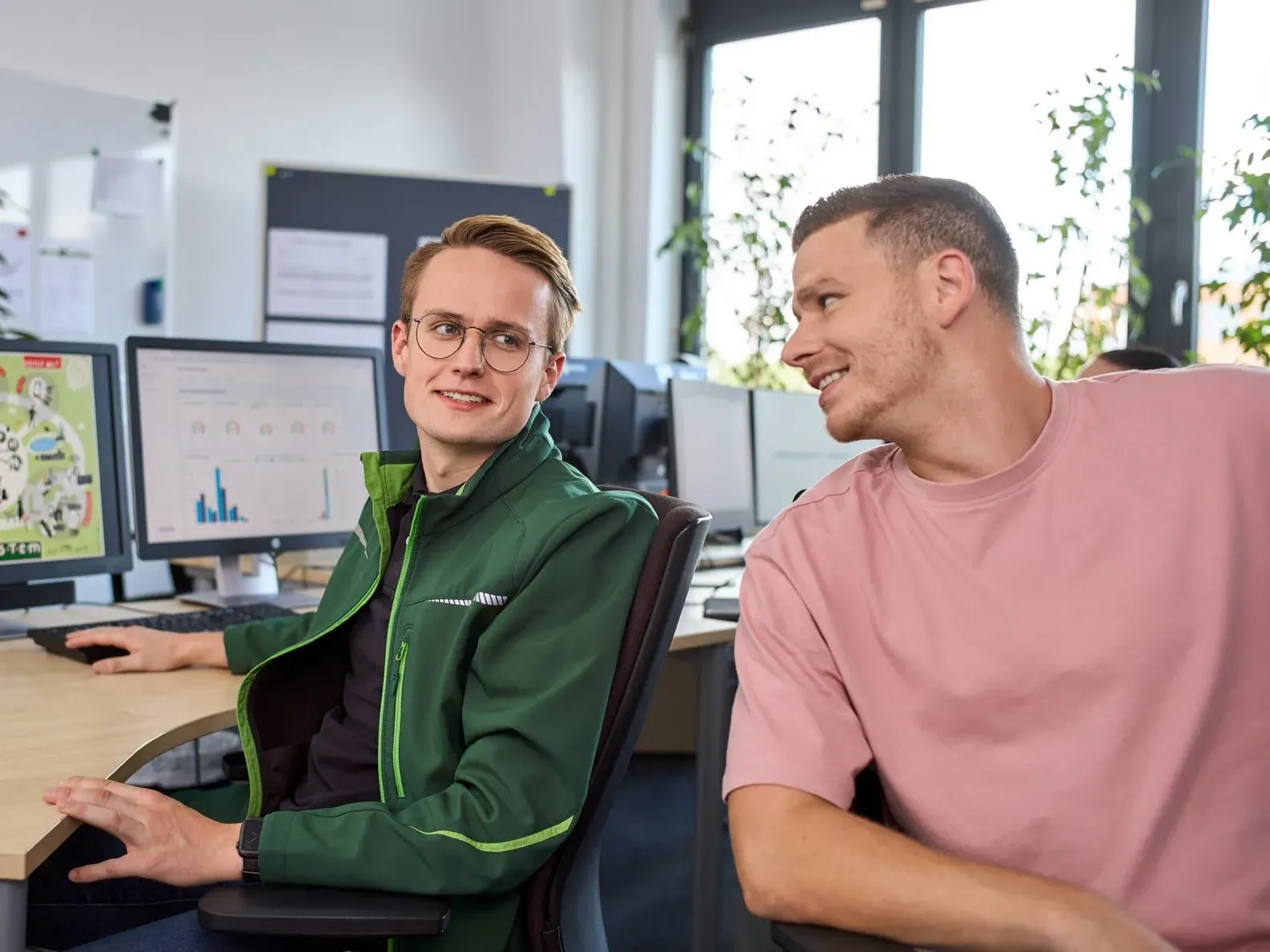 Zwei Kollegen in einem Büro, die sich unterhalten. Einer zeigt auf einen Bildschirm mit Diagrammen.