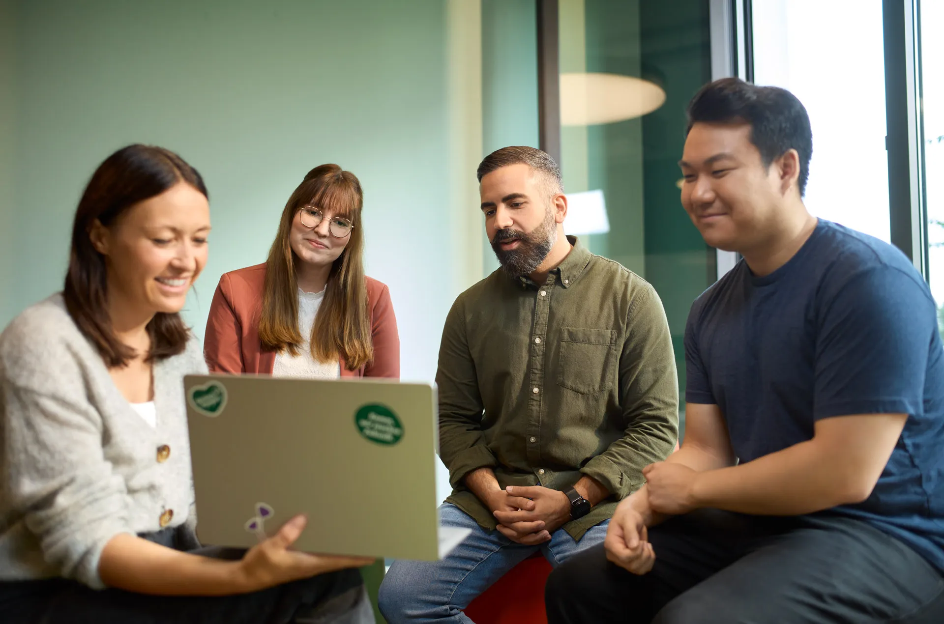 Eine Gruppe von vier Personen sitzt zusammen und schaut lächelnd auf einen Laptop in einem modernen Büro.