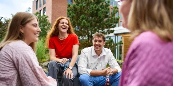 Vier junge Erwachsene sitzen lachend im Freien vor einem Bürogebäude und unterhalten sich in entspannter Atmosphäre.