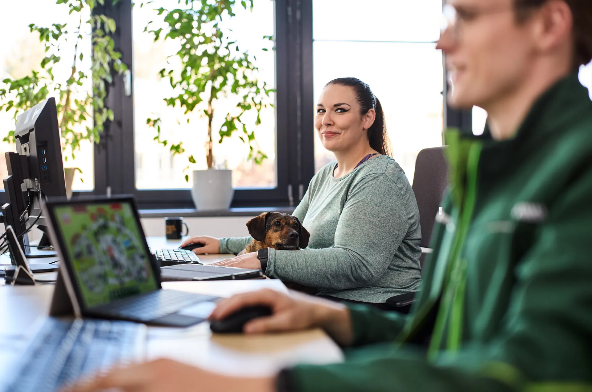 Eine Mitarbeiterin bei Fressnapf sitzt lächelnd am Schreibtisch mit einem Hund auf dem Schoß, während ein Kollege am Laptop arbeitet.