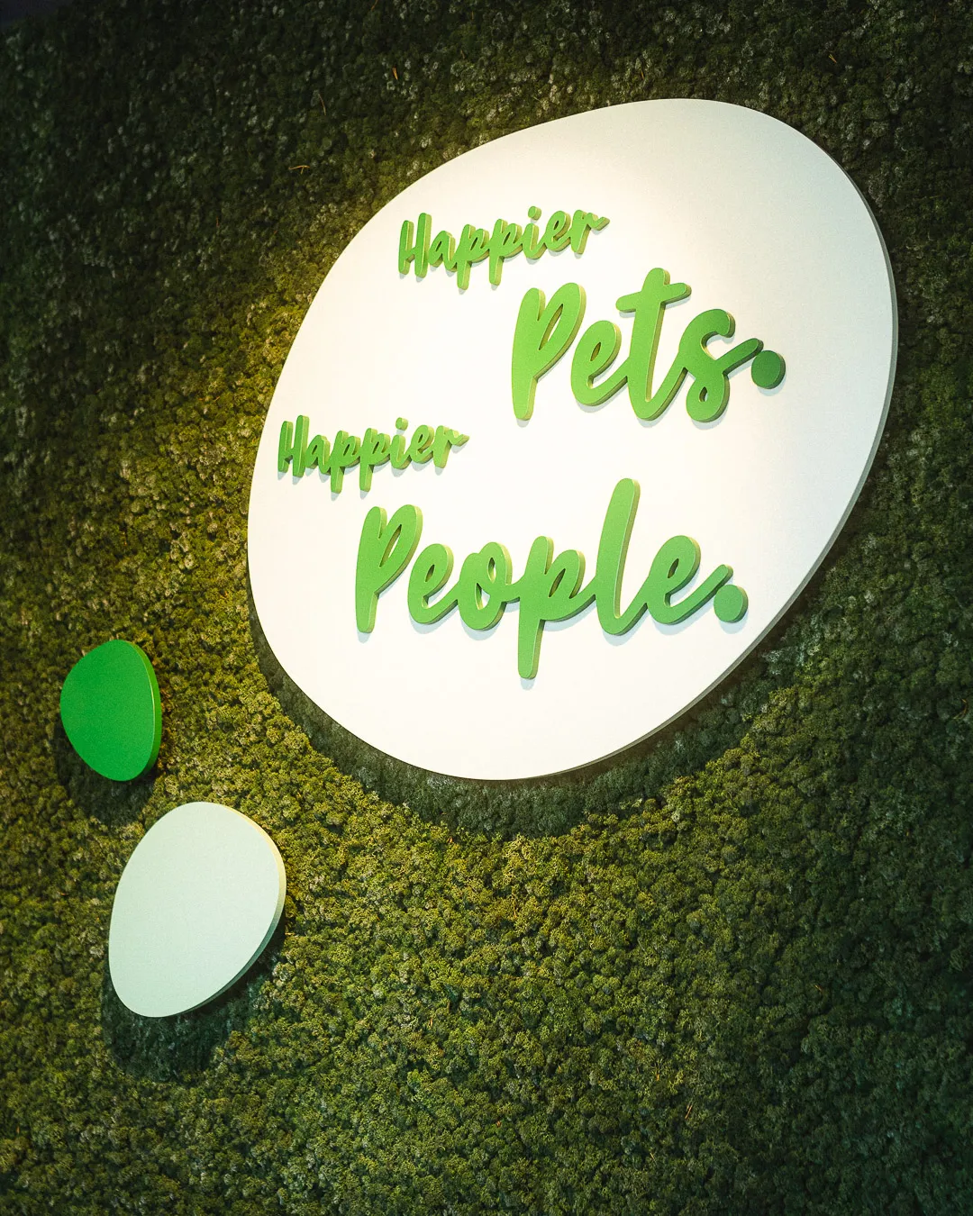 Zöld mohafalon “Happier Pets. Happier People.” felirat, barátságos és természetes hangulatban.